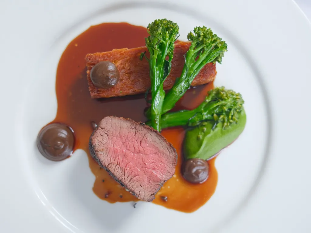 Plated fine dining dish with sliced pink beef, tenderstem broccoli, a crisp rectangular side and rich brown sauce on a white plate