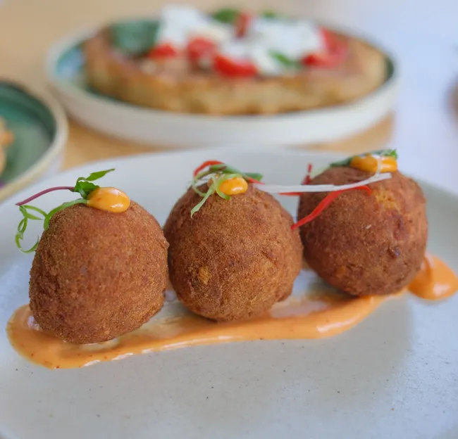 Three golden-brown fried croquettes served on a white plate with orange sauce and microgreens, with blurred brunch dishes in the background.