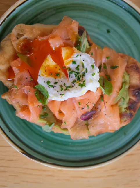 Overhead view of brunch dishes on a wooden table, including smoked salmon with a poached egg on flatbread, a tomato-topped flatbread, and a bowl of fritters garnished with shredded vegetables