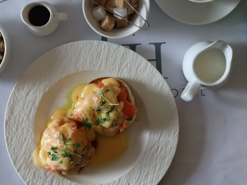Overhead view of eggs Benedict on a white plate with coffee, milk and a bowl of sugar cubes on the table