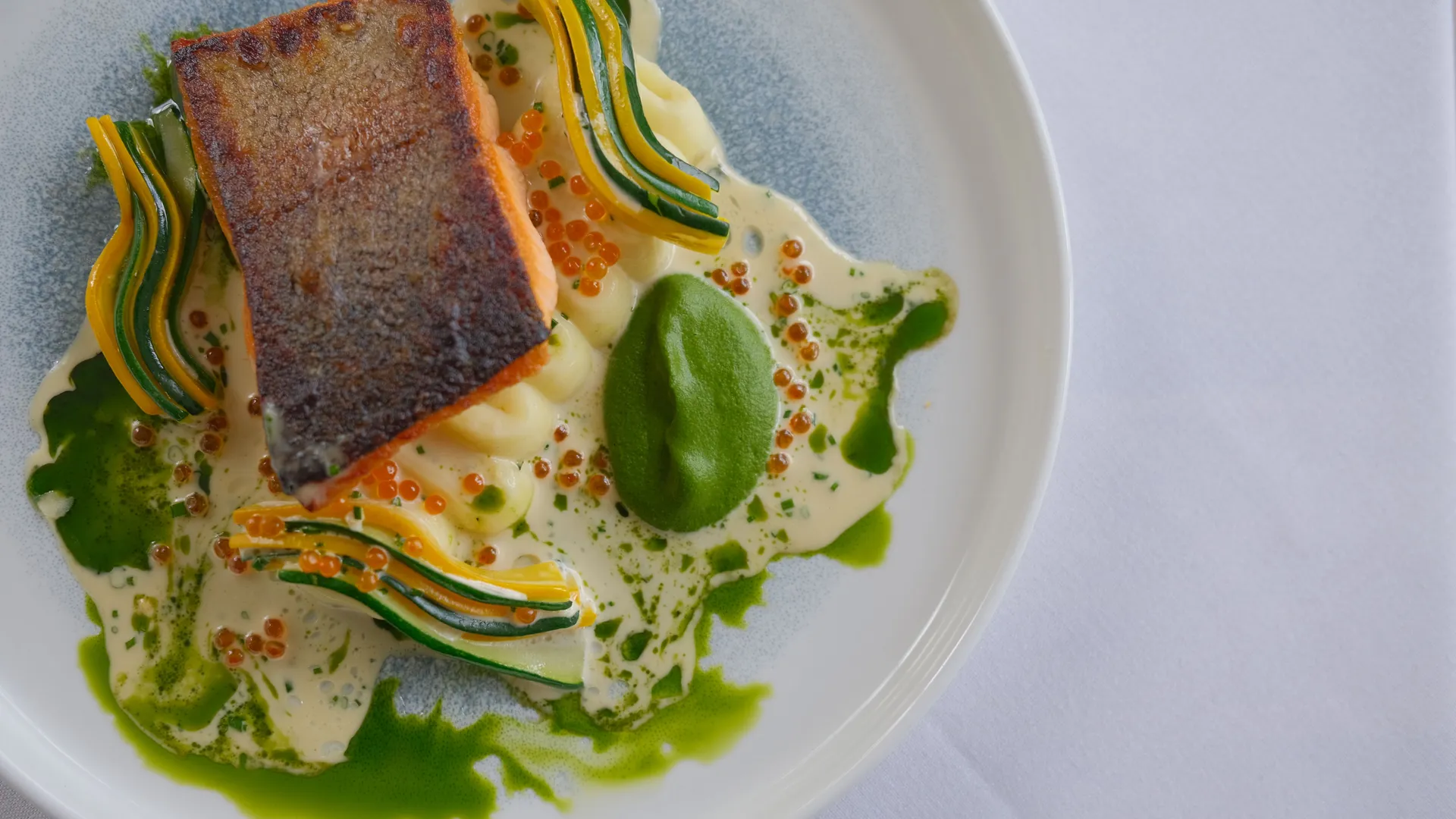 Pan-fried salmon fillet with crisp skin, piped purée, ribboned courgette and yellow squash, green herb sauce and salmon roe on a white plate