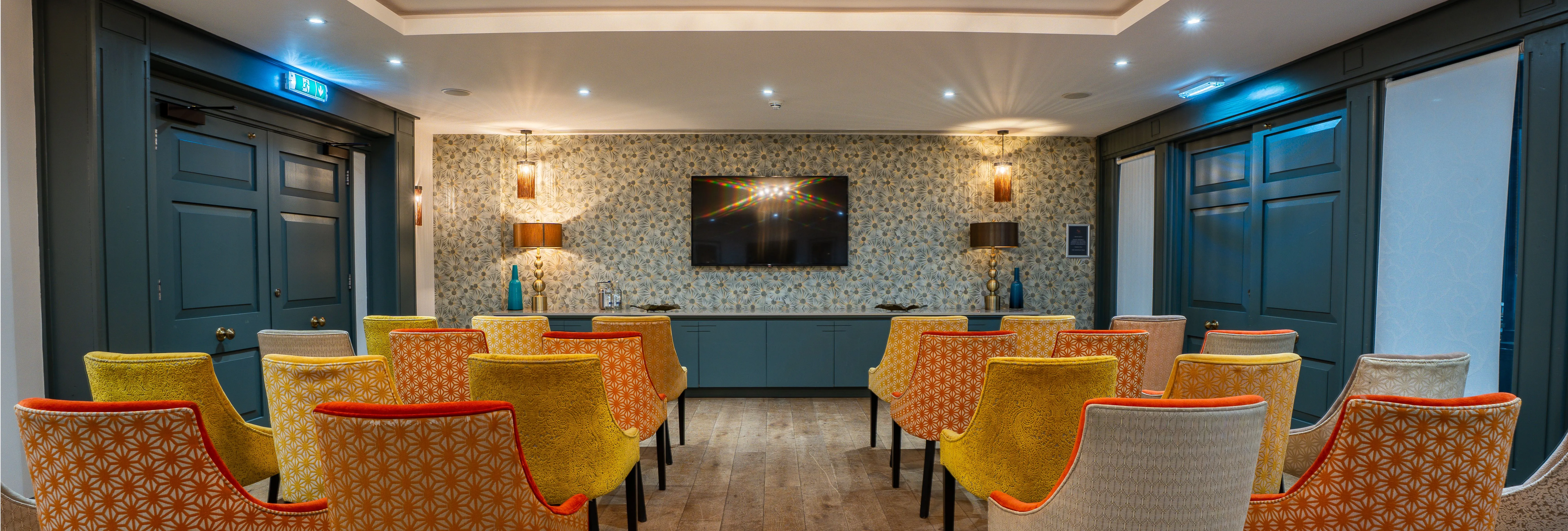 Stylish meeting or presentation room with patterned upholstered chairs, dark blue panelling, wooden flooring and a wall-mounted television.