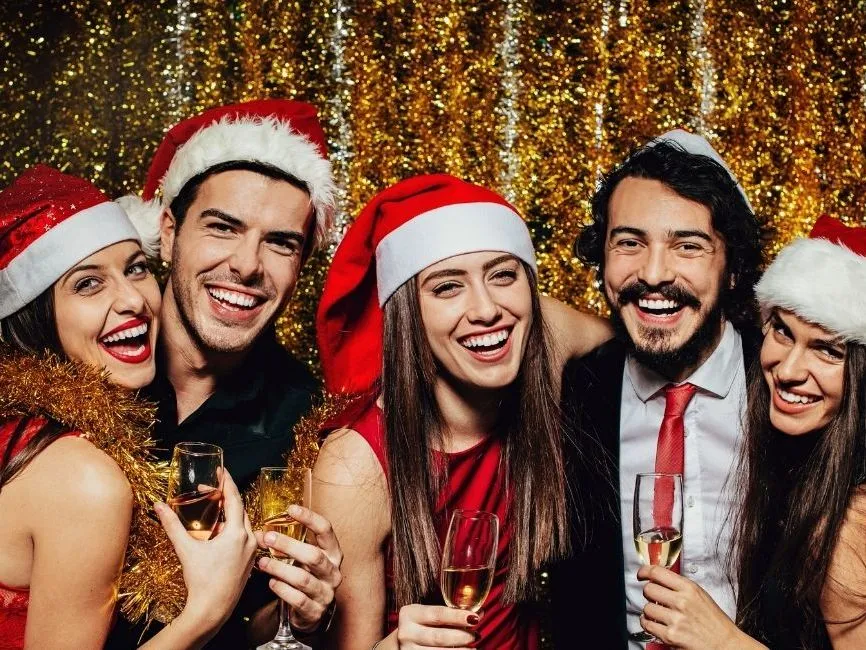 Five friends in Santa hats laughing and holding champagne glasses before a gold tinsel backdrop.