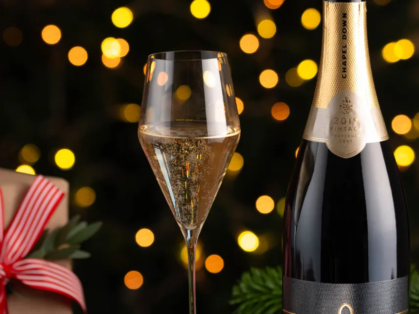 Bottle of Chapel Down sparkling wine beside a filled champagne flute and wrapped gift, with warm festive lights in the background.