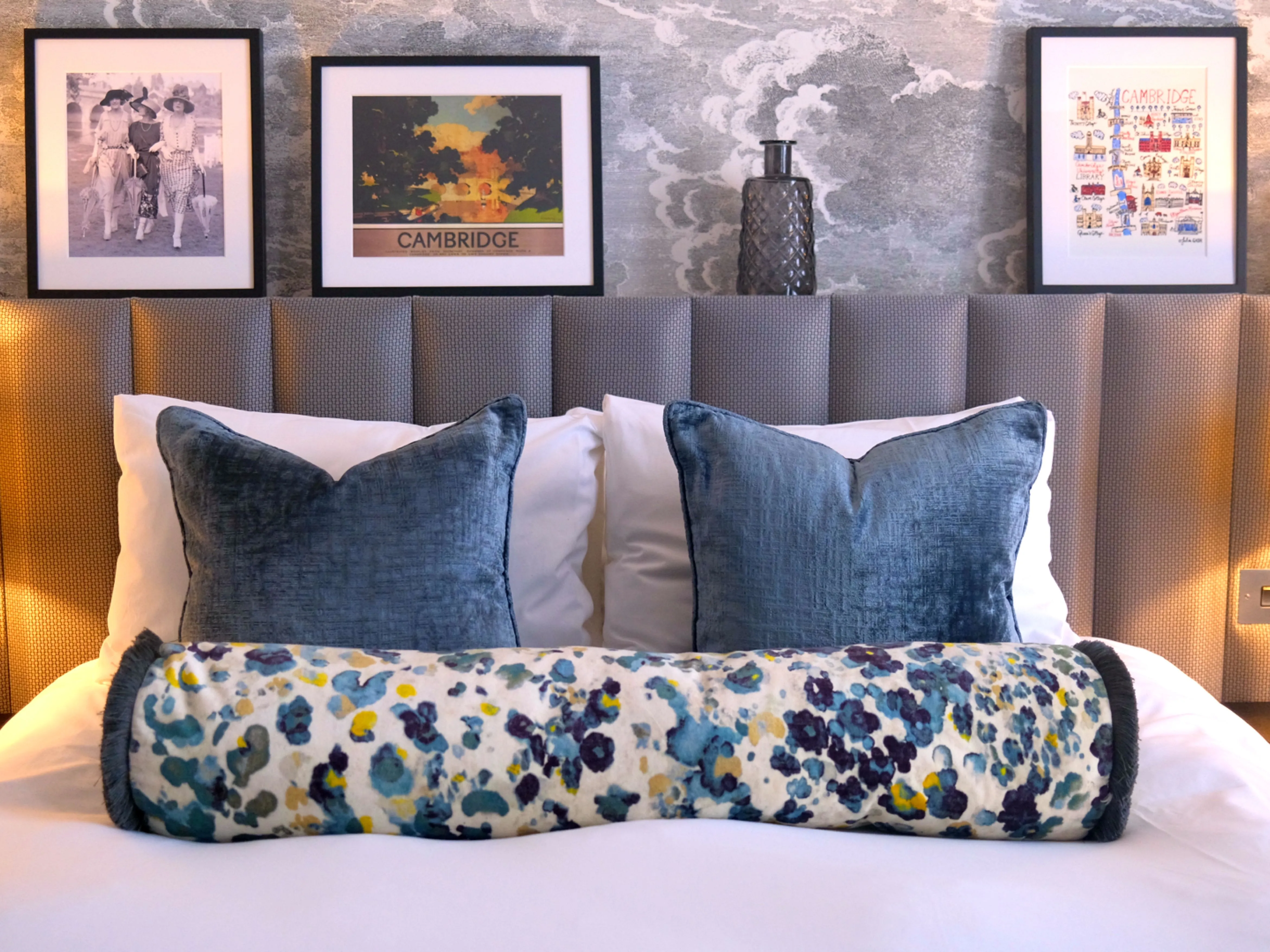 Cosy bedroom with a cushioned headboard, blue velvet cushions, floral bolster and warm bedside lamps.