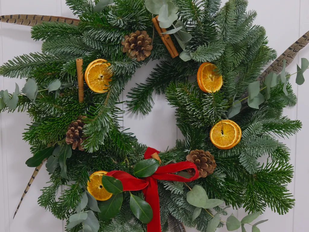 Festive evergreen wreath with dried orange slices, pinecones, cinnamon sticks, feathers and a red ribbon on a white door.