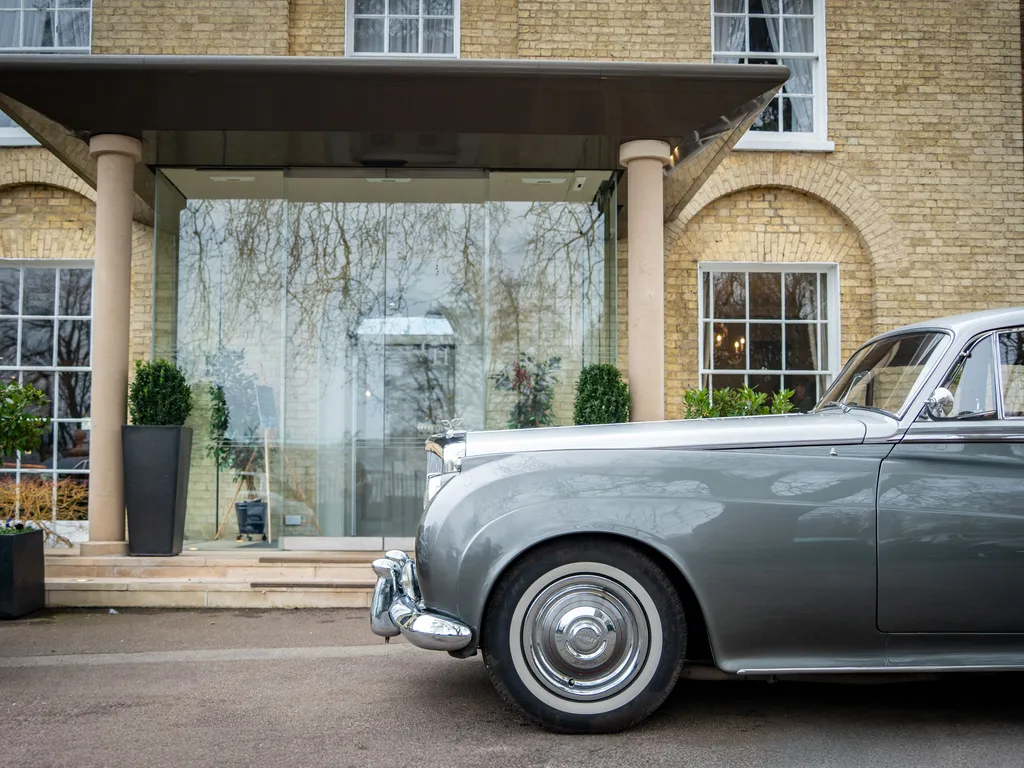 Classic silver luxury car parked outside a brick building with a covered glass entrance