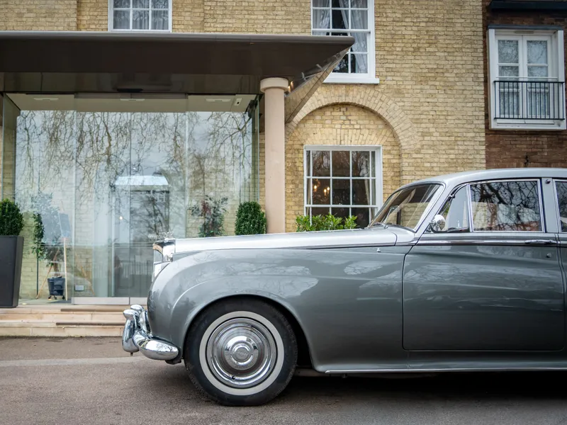 Classic silver luxury car parked outside a brick building with a covered glass entrance