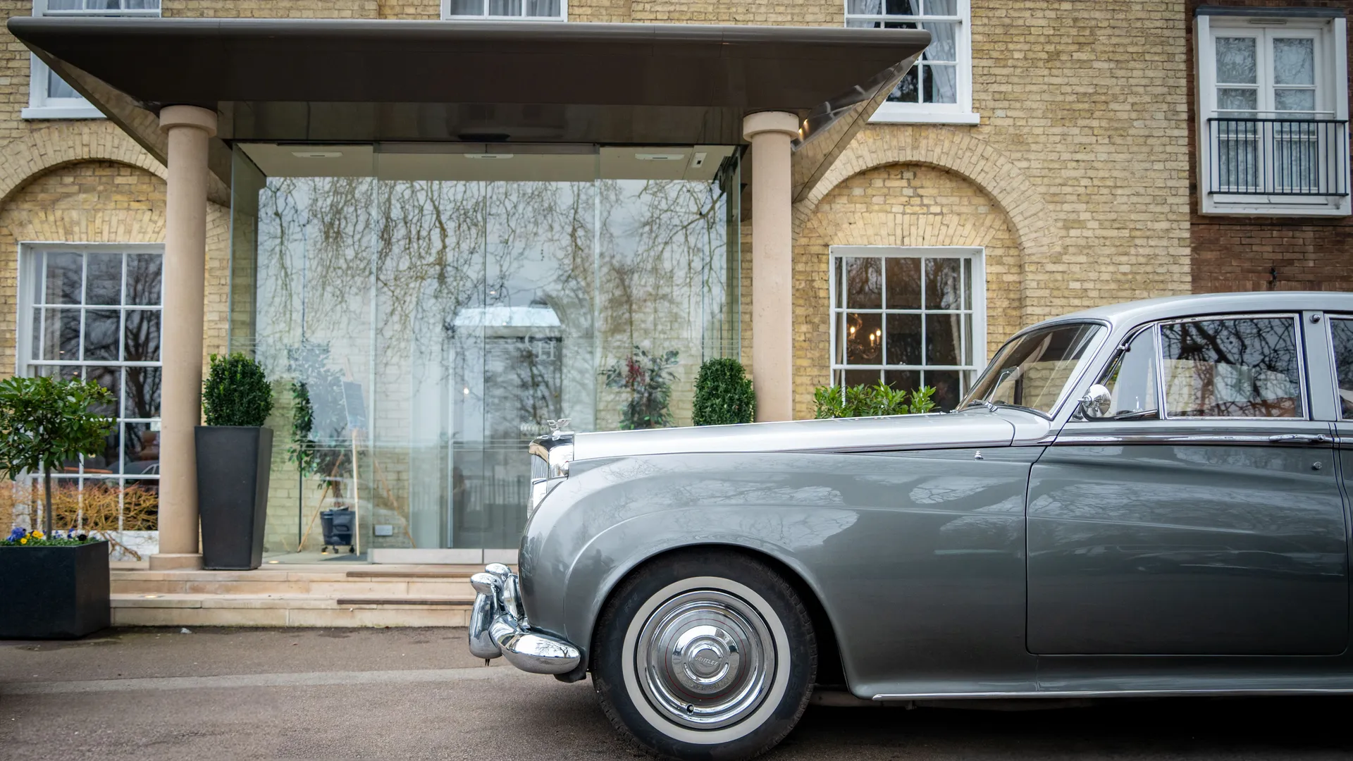 Classic silver luxury car parked outside a brick building with a covered glass entrance
