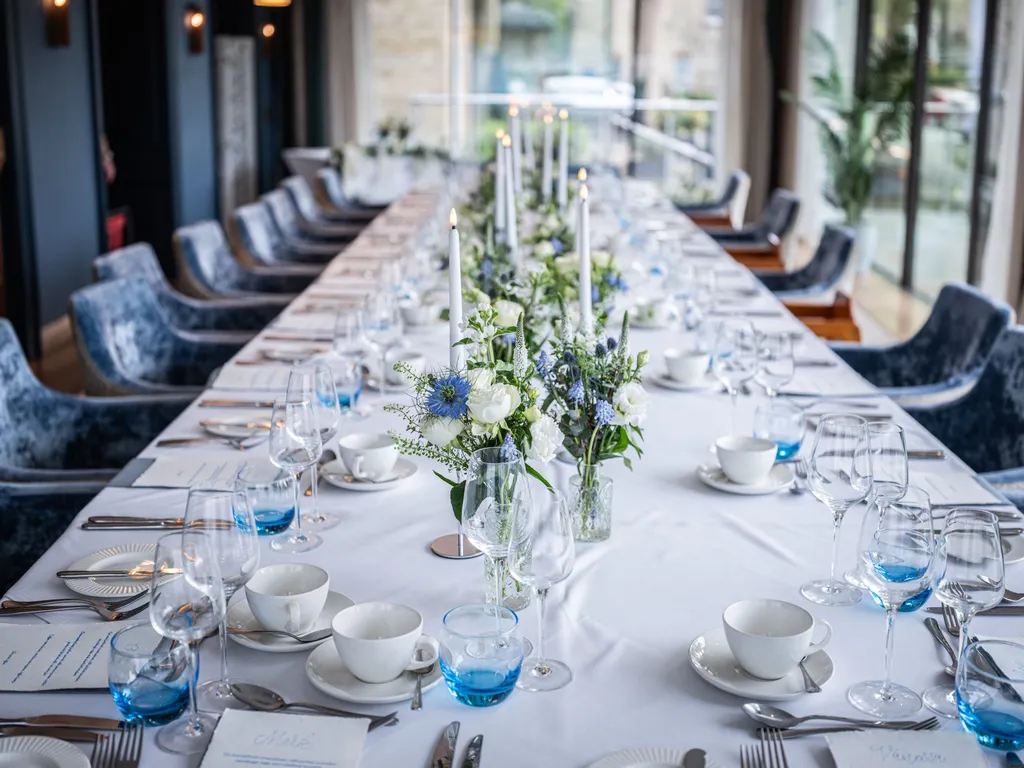 Elegant long dining table set with blue velvet chairs, white and blue flowers, candles and glassware