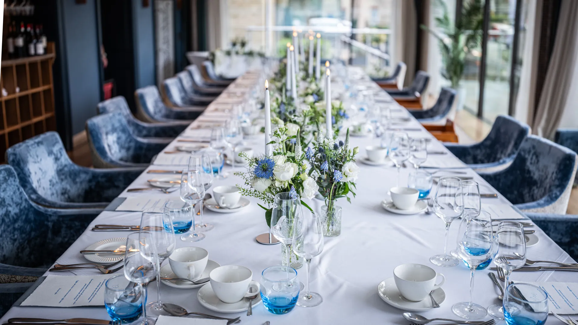 Elegant long dining table set with blue velvet chairs, white and blue flowers, candles and glassware