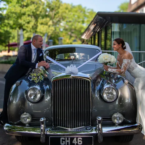 Bride and groom smiling at each other across the bonnet of a ribbon-decorated vintage wedding car