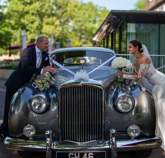 Bride and groom smiling at each other across the bonnet of a ribbon-decorated vintage wedding car