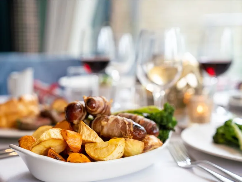 Bowl of roast potatoes and bacon-wrapped sausages with greens on a restaurant table