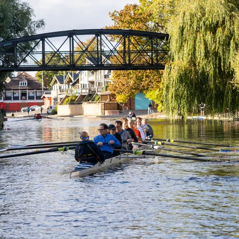 Rowing crew in a long boat on a tree-lined river beneath a black footbridge