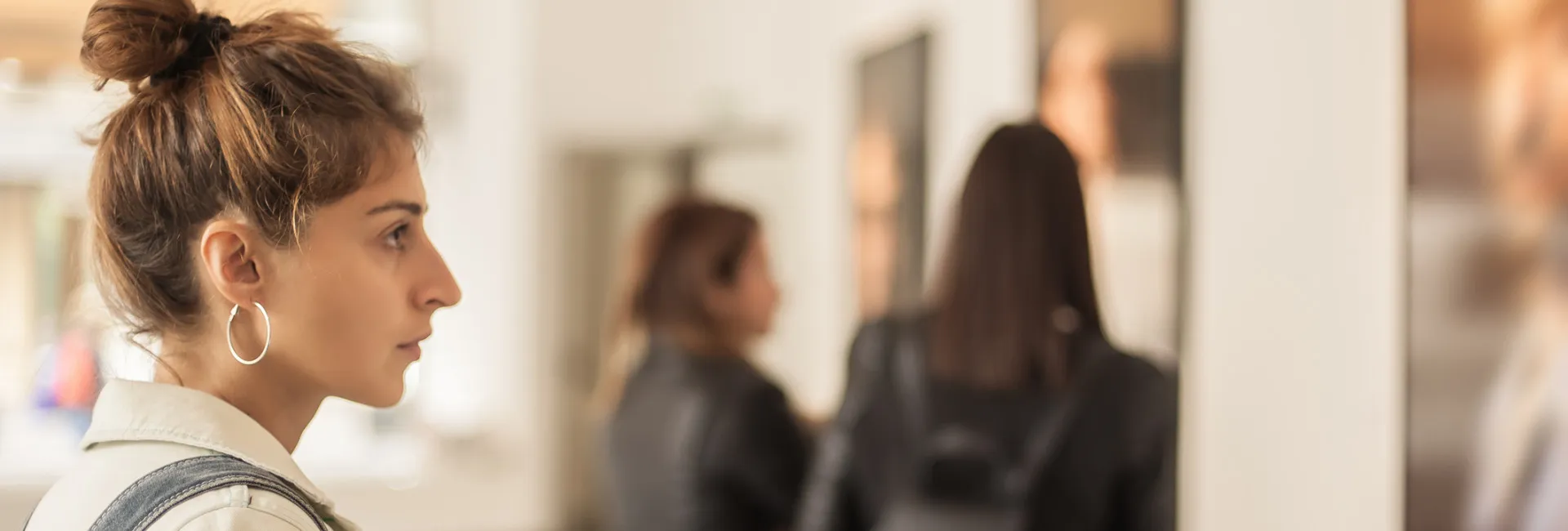 A woman with her hair in a messy bun and hoop earrings views art on display in a gallery, while two other women in dark jackets are seen blurred in the background.