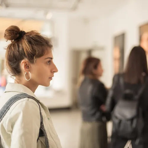 A woman with her hair in a messy bun and hoop earrings views art on display in a gallery, while two other women in dark jackets are seen blurred in the background.