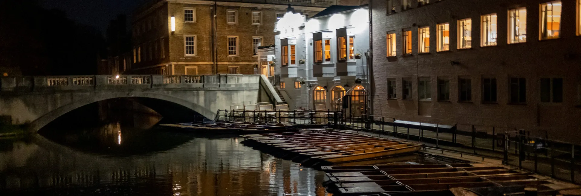 A nighttime view of a riverside scene with a stone bridge, illuminated buildings reflecting in the water, and a row of empty wooden boats moored along the bank under dim lighting.