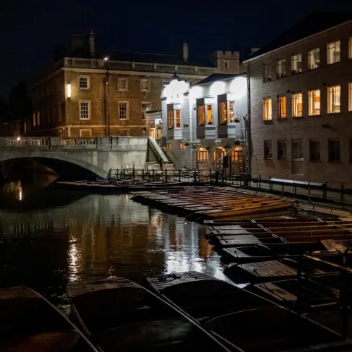 A nighttime view of a riverside scene with a stone bridge, illuminated buildings reflecting in the water, and a row of empty wooden boats moored along the bank under dim lighting.