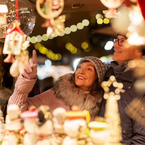 A smiling couple enjoys a Christmas market, admiring festive decorations. The scene is illuminated by warm string lights, with holiday ornaments and trinkets surrounding them.