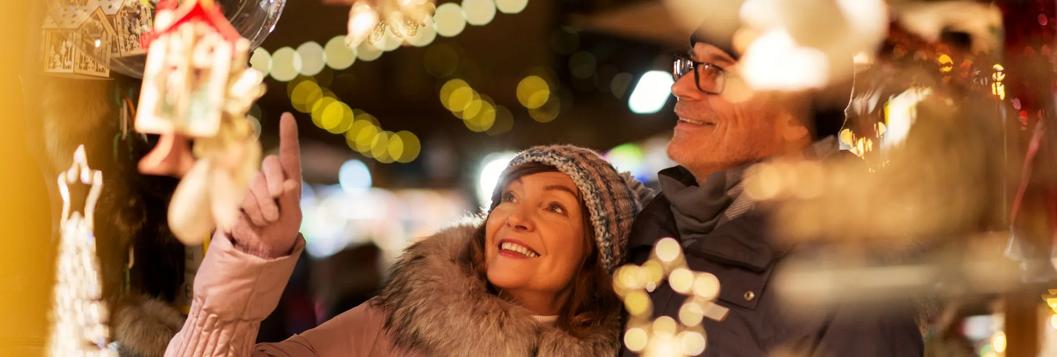 A smiling couple enjoys a Christmas market, admiring festive decorations. The scene is illuminated by warm string lights, with holiday ornaments and trinkets surrounding them.
