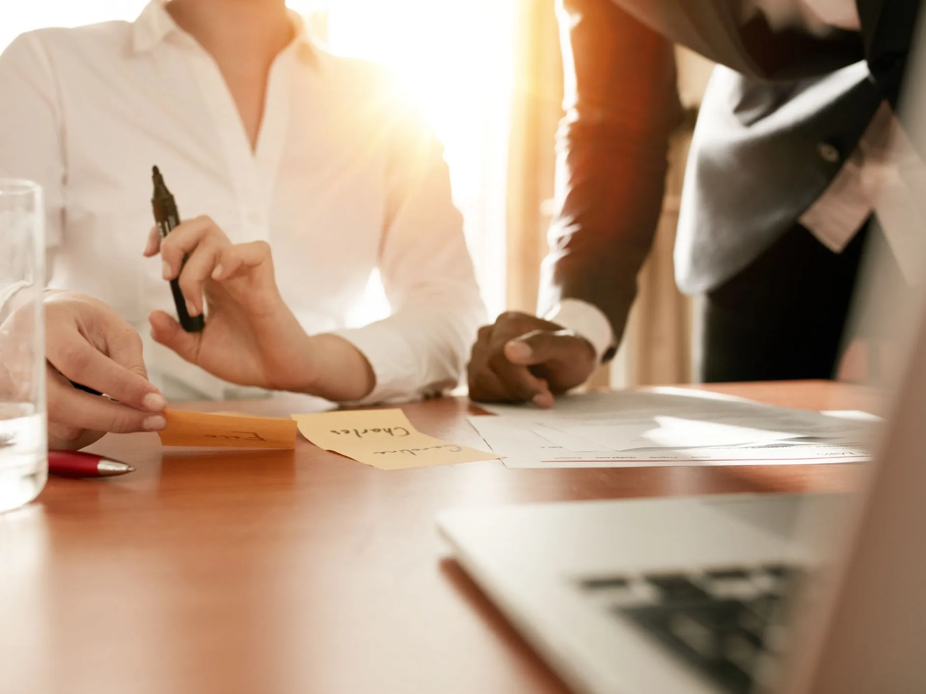 Two individuals in a business setting collaborate over documents and sticky notes on a table, with warm sunlight streaming in through a window. One person holds a marker.