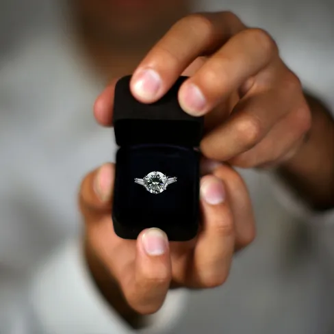 A close-up of hands holding an open black ring box with a sparkling diamond engagement ring, set against a blurred background.