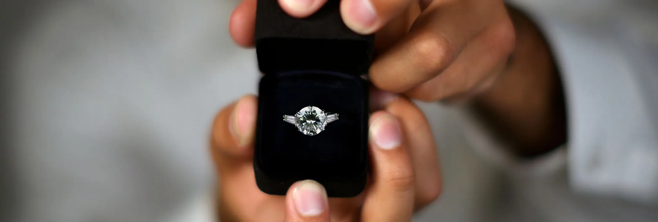A close-up of hands holding an open black ring box with a sparkling diamond engagement ring, set against a blurred background.