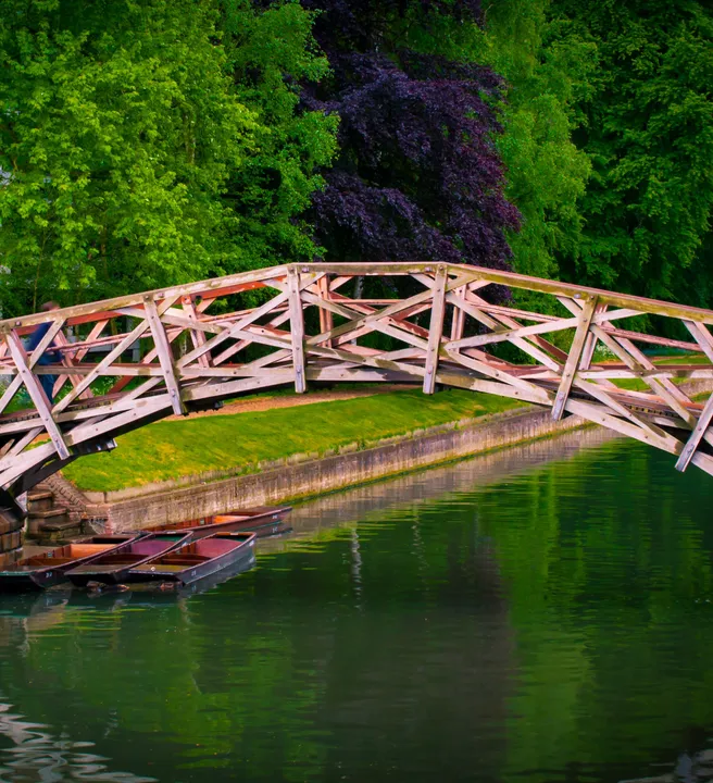 A wooden bridge with intricate crisscross patterns spans a calm river surrounded by lush green trees, with small boats docked beneath and a historic brick building nearby.