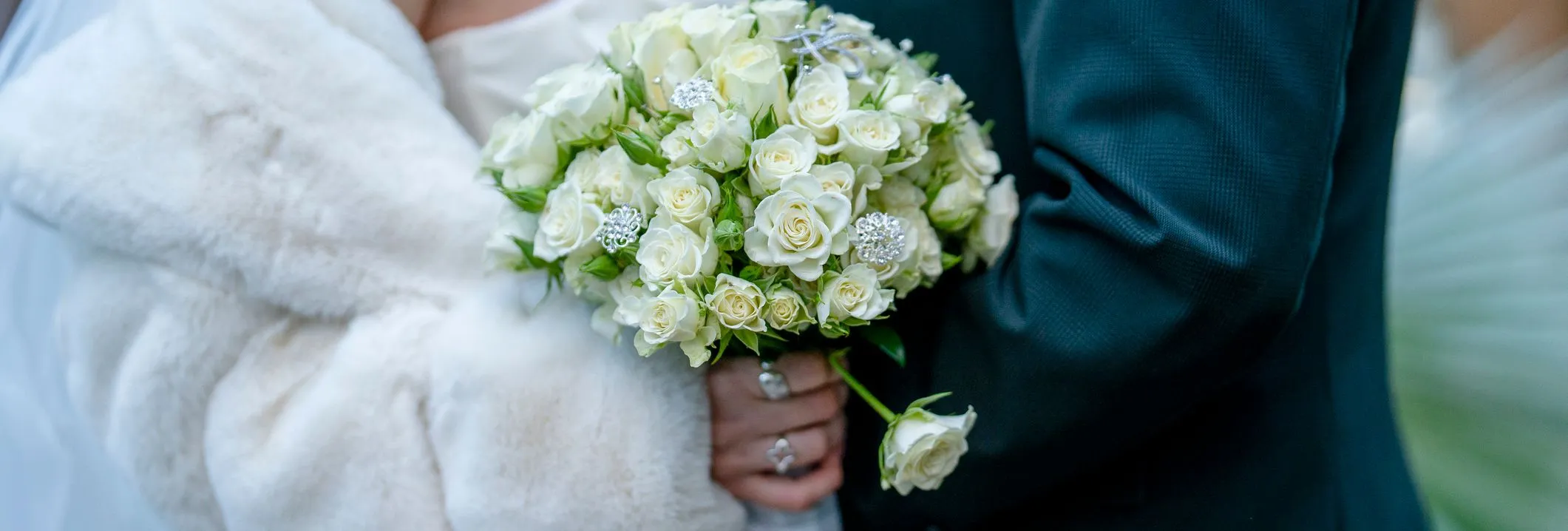 A close-up of a bride in a white fur wrap holding a bouquet of white roses with decorative accents, standing next to a groom in a dark suit.