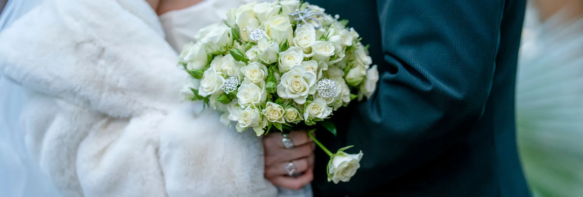 A close-up of a bride in a white fur wrap holding a bouquet of white roses with decorative accents, standing next to a groom in a dark suit.