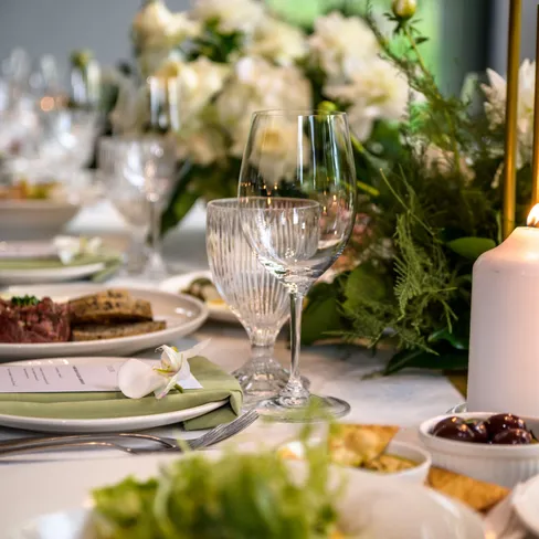 A beautifully set dining table with elegant glassware, green napkins, white plates, lit candles, and lush floral arrangements featuring white and green accents.