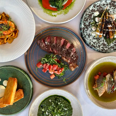 A top-down view of a gourmet meal with various plated dishes, including steak, pasta with shrimp, crispy potatoes, fish, and colorful vegetable sides.