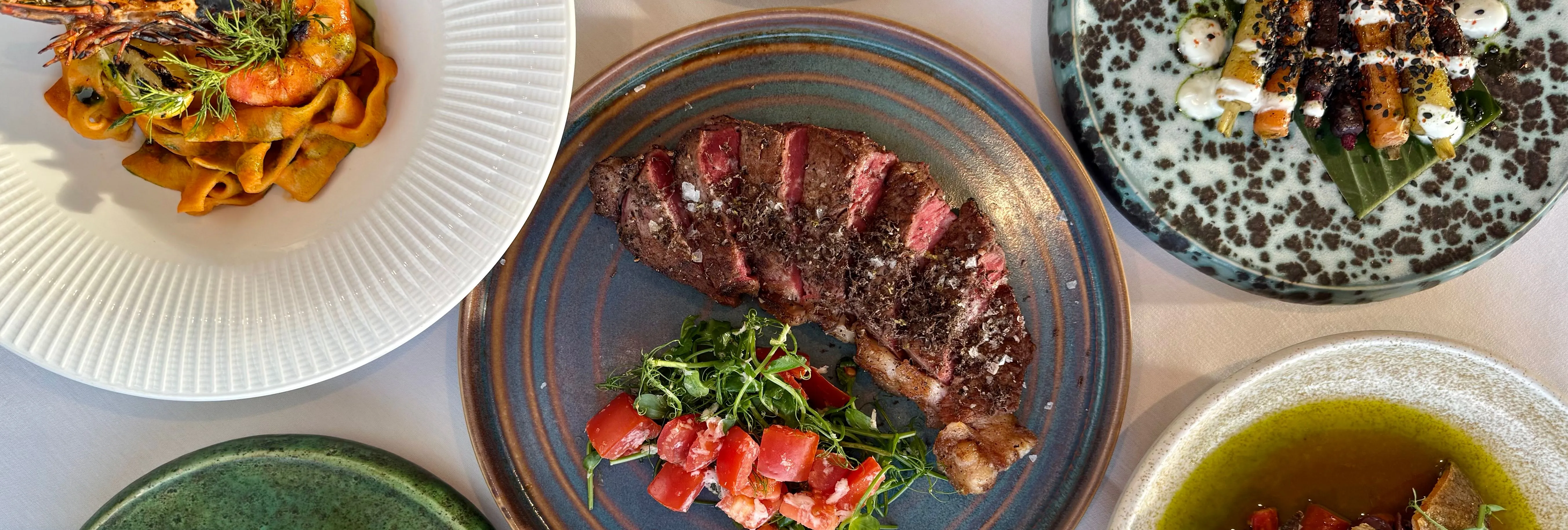 A top-down view of a gourmet meal with various plated dishes, including steak, pasta with shrimp, crispy potatoes, fish, and colorful vegetable sides.