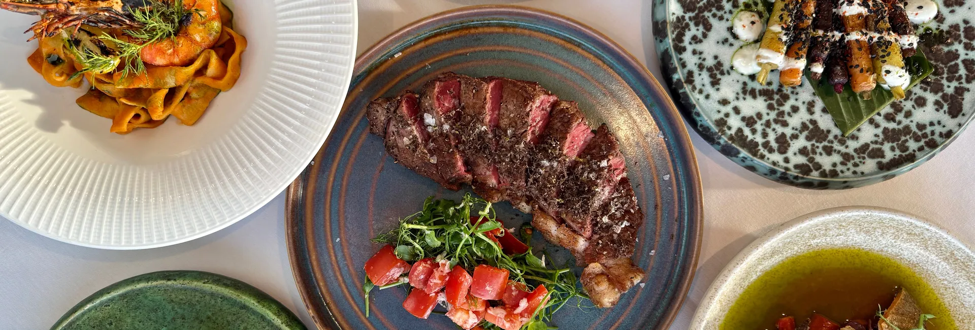 A top-down view of a gourmet meal with various plated dishes, including steak, pasta with shrimp, crispy potatoes, fish, and colorful vegetable sides.