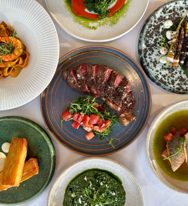 A top-down view of a gourmet meal with various plated dishes, including steak, pasta with shrimp, crispy potatoes, fish, and colorful vegetable sides.