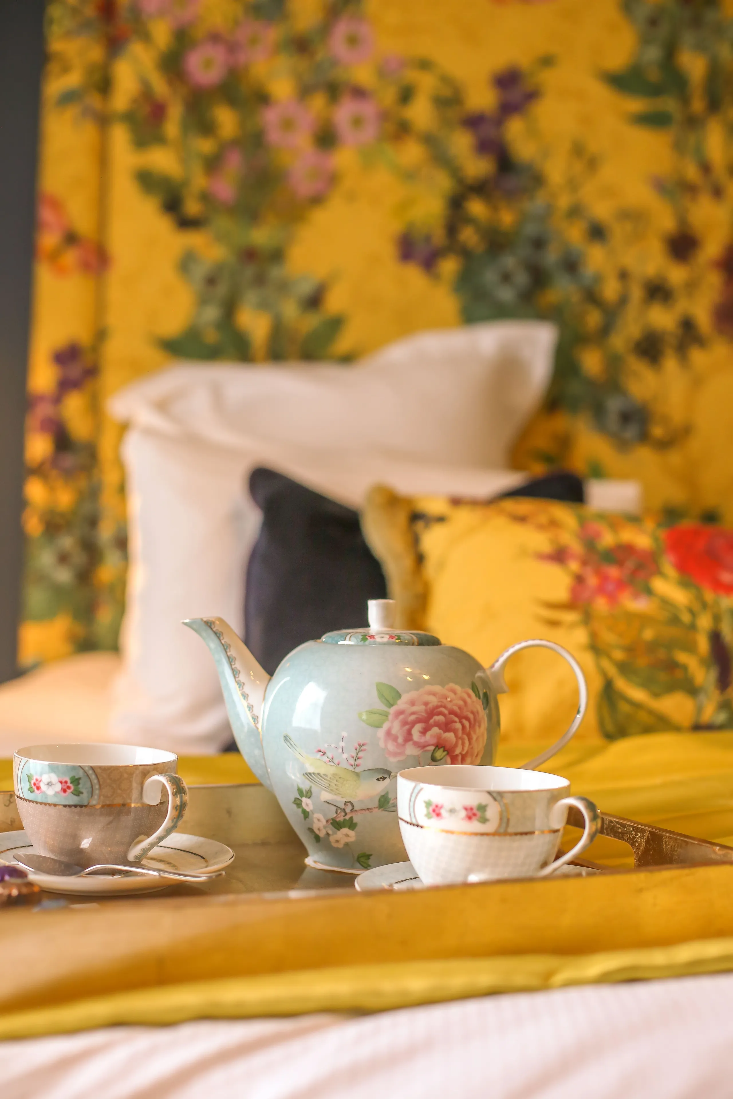A floral teapot and two teacups on a tray atop the bed in the Rosa feature room at Gonville Hotel, with vibrant cushions and a floral headboard.