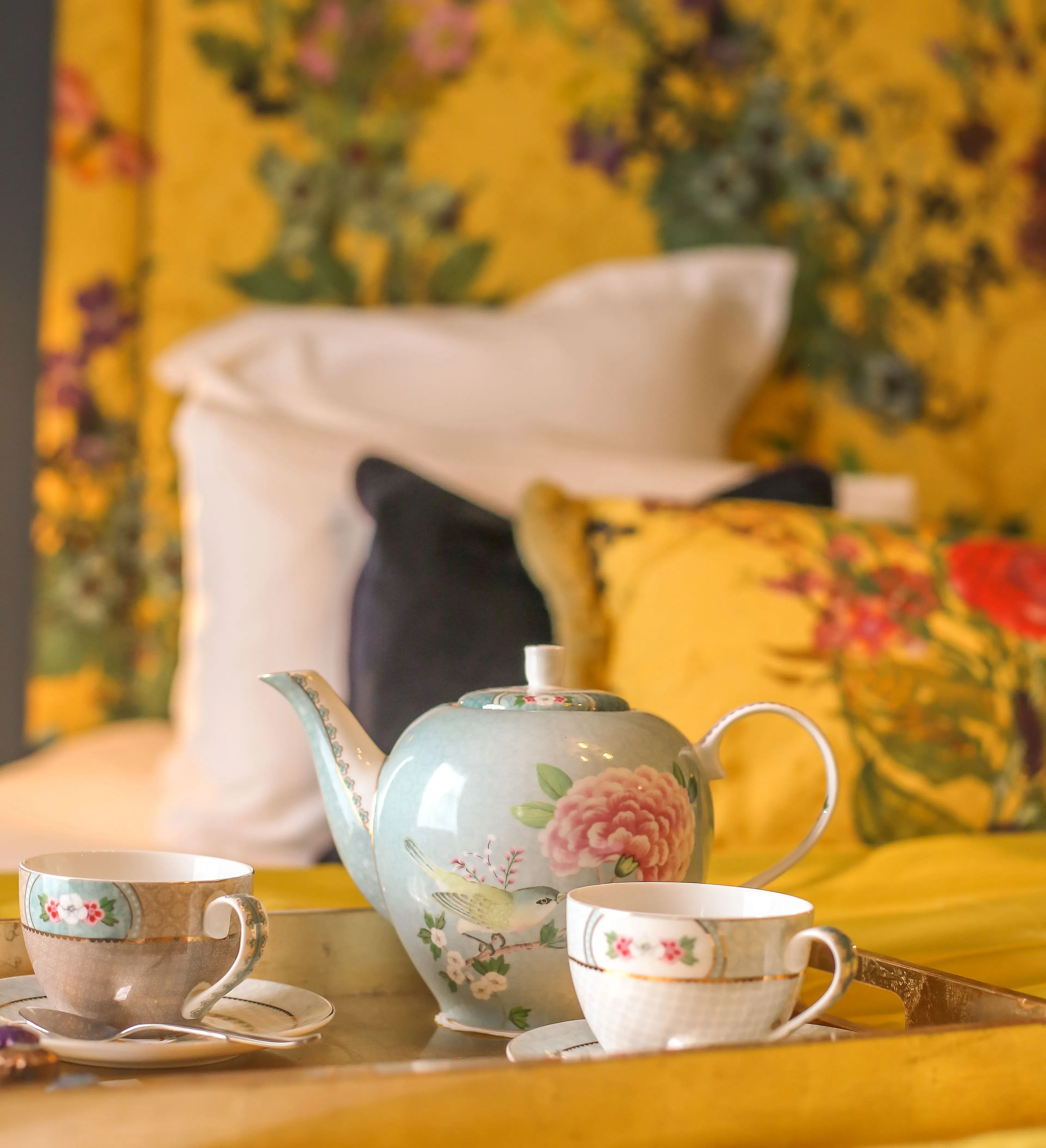 A floral teapot and two teacups on a tray atop the bed in the Rosa feature room at Gonville Hotel, with vibrant cushions and a floral headboard.