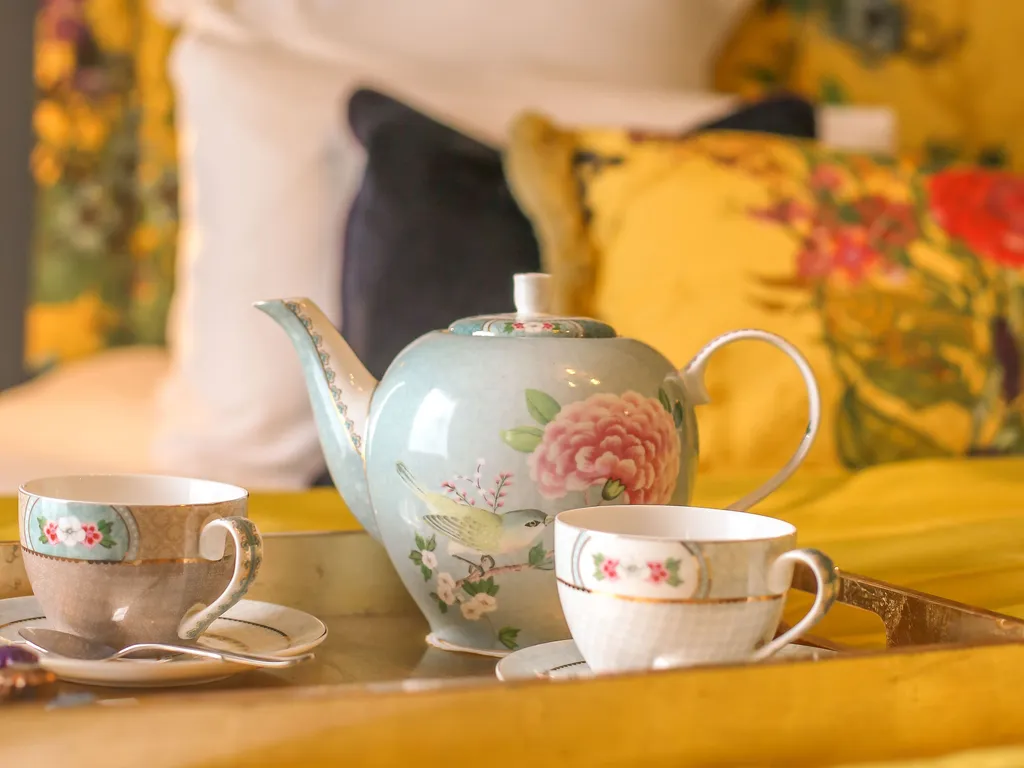 A floral teapot and two teacups on a tray atop the bed in the Rosa feature room at Gonville Hotel, with vibrant cushions and a floral headboard.