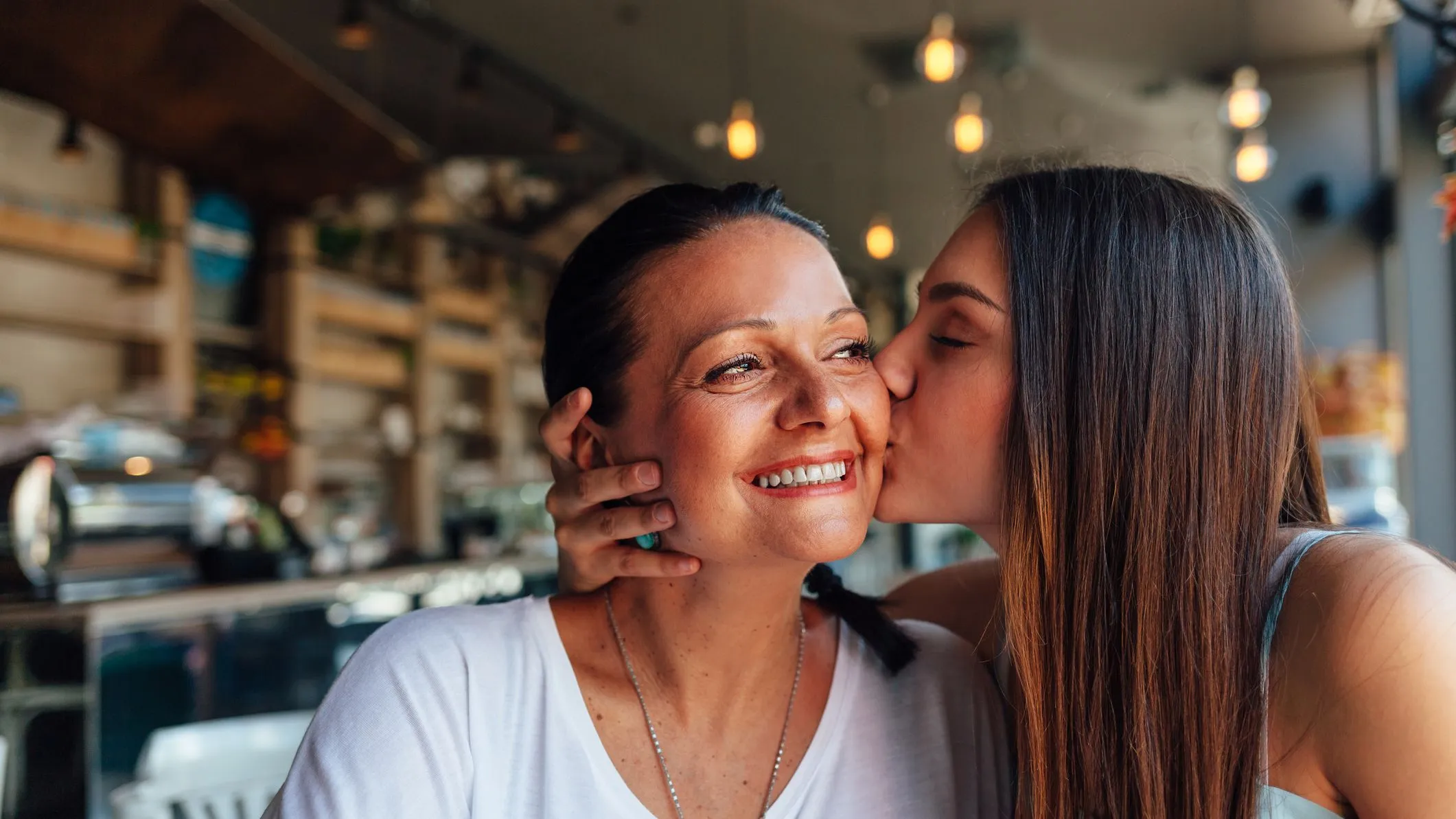 A young woman kisses an older woman on the cheek in a warmly lit café. The older woman smiles joyfully, her eyes crinkling with happiness.