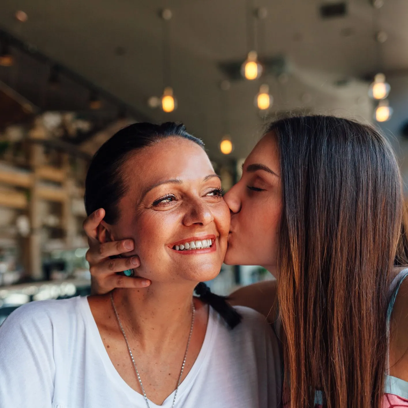 A young woman kisses an older woman on the cheek in a warmly lit café. The older woman smiles joyfully, her eyes crinkling with happiness.