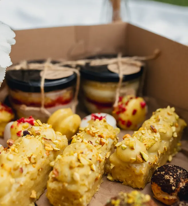 A box of assorted desserts, including pistachio-topped pastries, chocolate-covered treats, and jarred layered cakes, tied with twine, set on brown parchment paper.