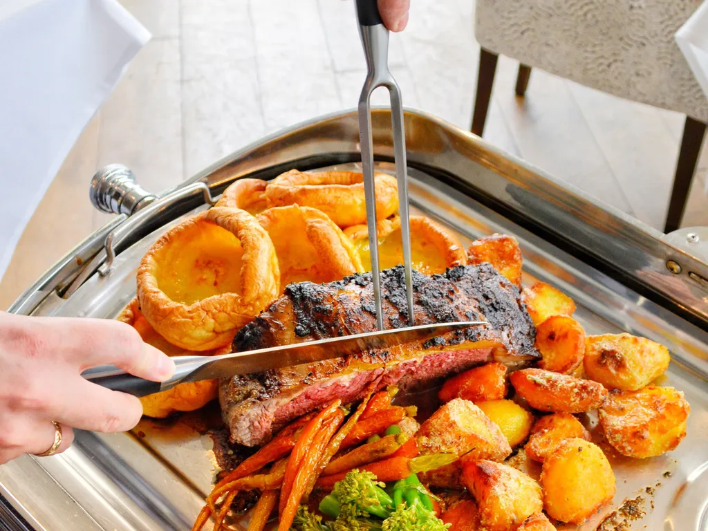 A person carves a medium-rare roast beef on a silver tray, surrounded by golden roasted potatoes, Yorkshire puddings, glazed carrots, and green broccolini.
