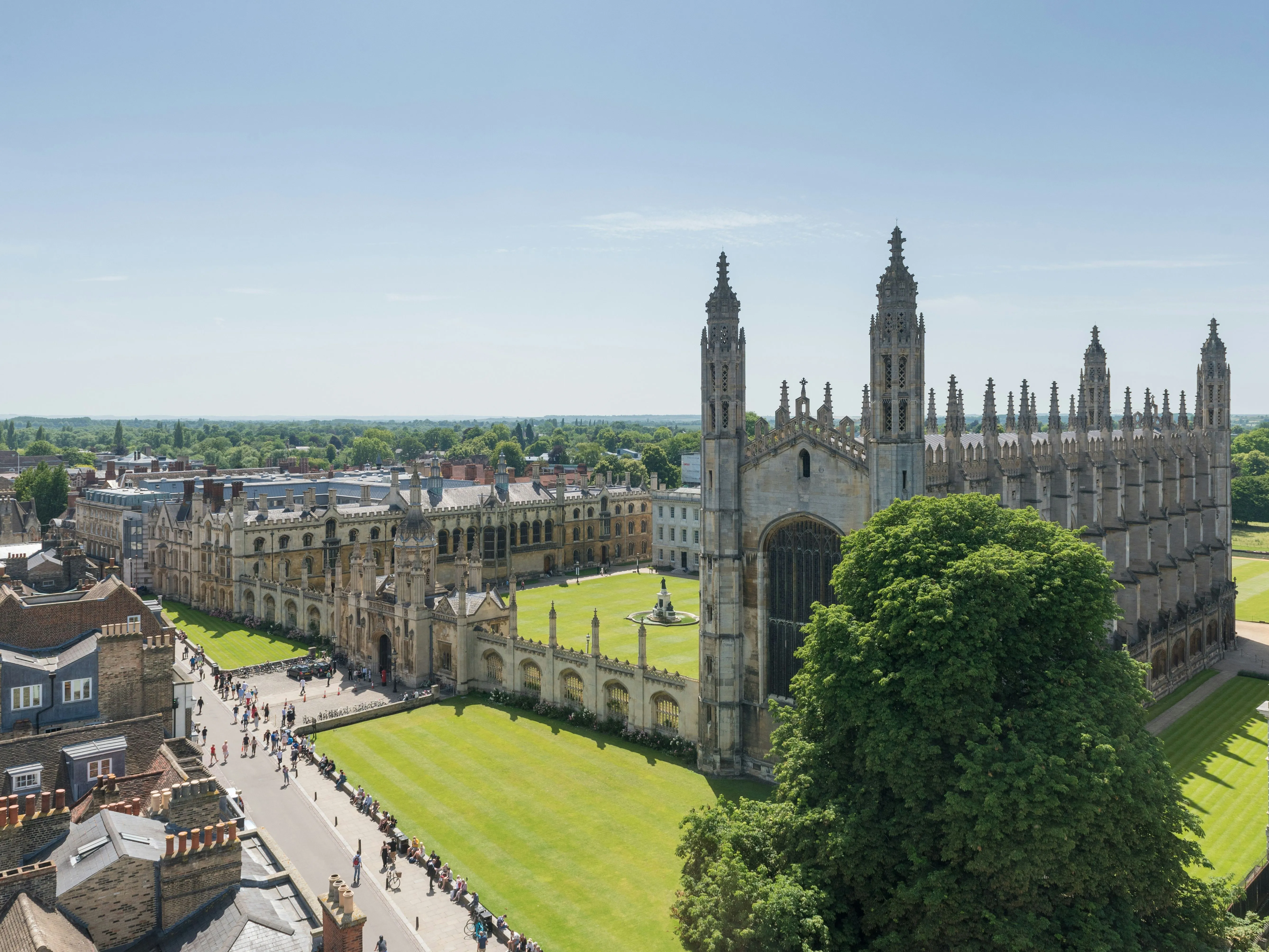 Aerial view of King's College Chapel and its surrounding courtyard in Cambridge, with tourists walking along nearby paths on a bright, sunny day.