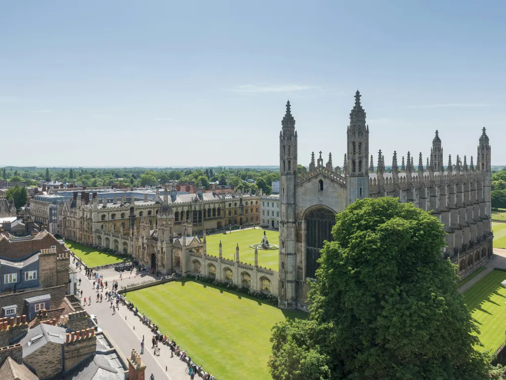 Aerial view of King's College Chapel and its surrounding courtyard in Cambridge, with tourists walking along nearby paths on a bright, sunny day.