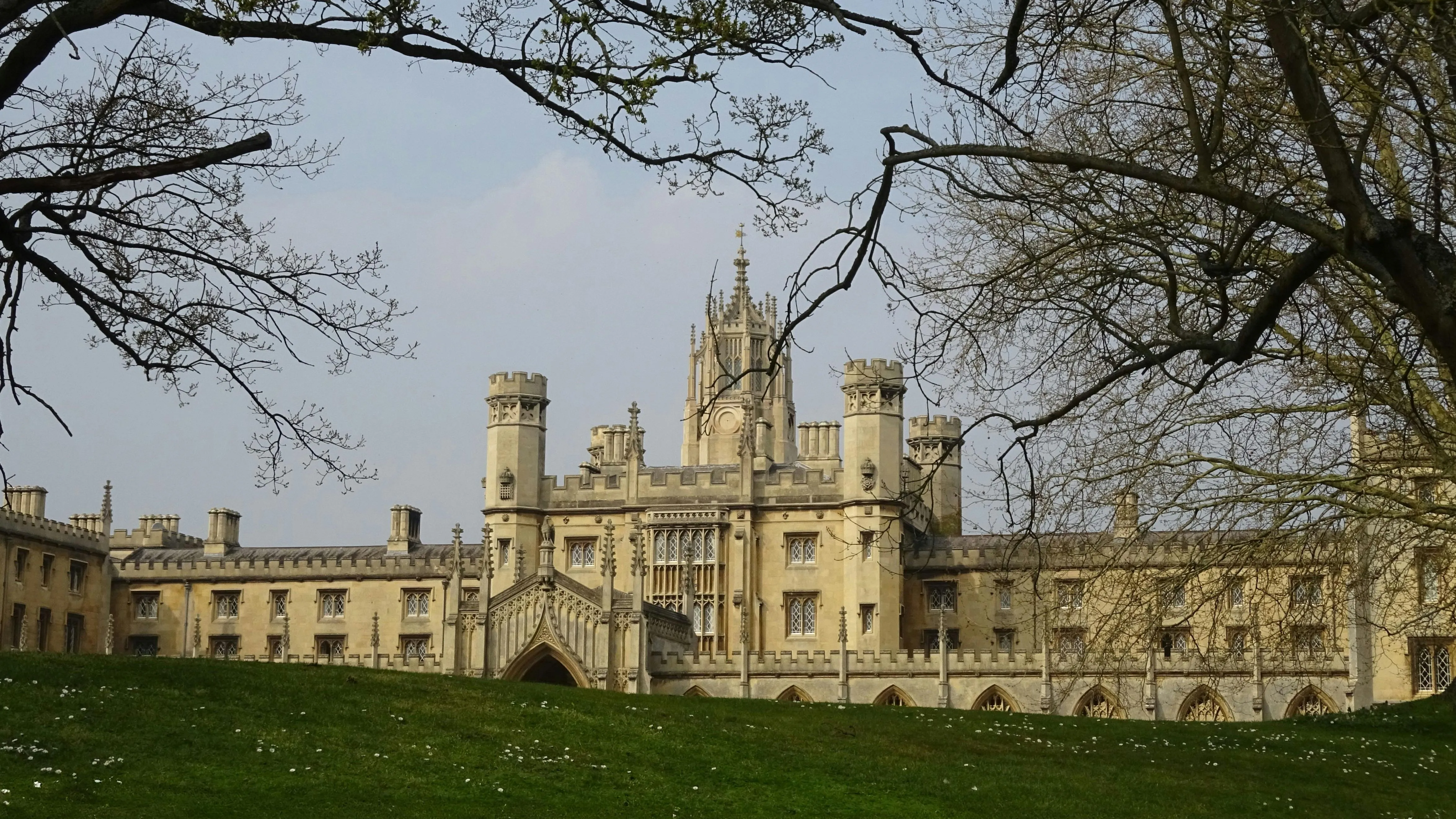 Historic Gothic-style college building with tall towers and arched windows, framed by leafless trees and a grassy lawn under a pale blue sky.