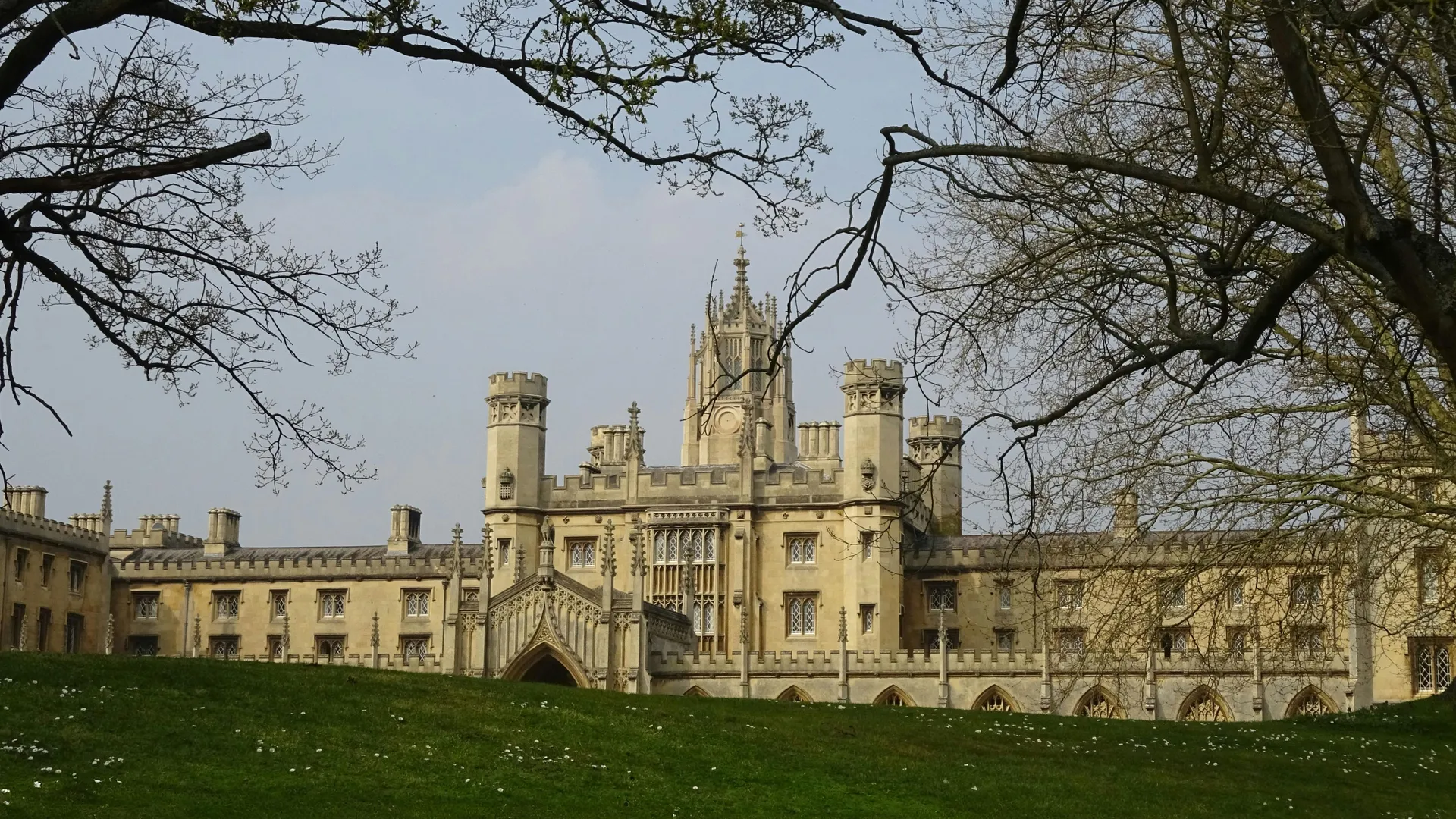 Historic Gothic-style college building with tall towers and arched windows, framed by leafless trees and a grassy lawn under a pale blue sky.
