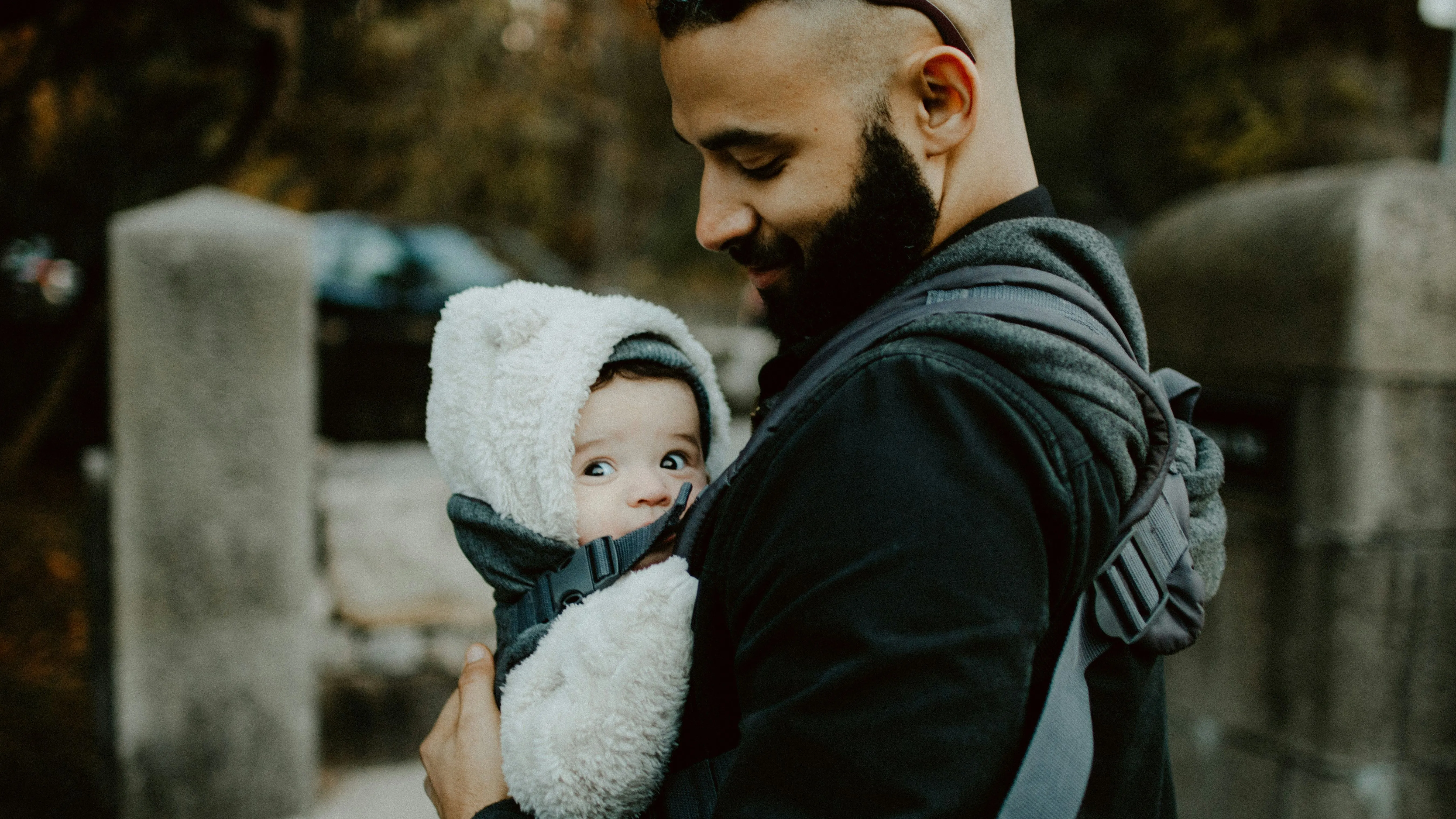 Smiling man with a beard carrying a bundled-up baby in a front carrier on a chilly day; the baby looks to the side while snuggled in a fluffy hooded outfit.
