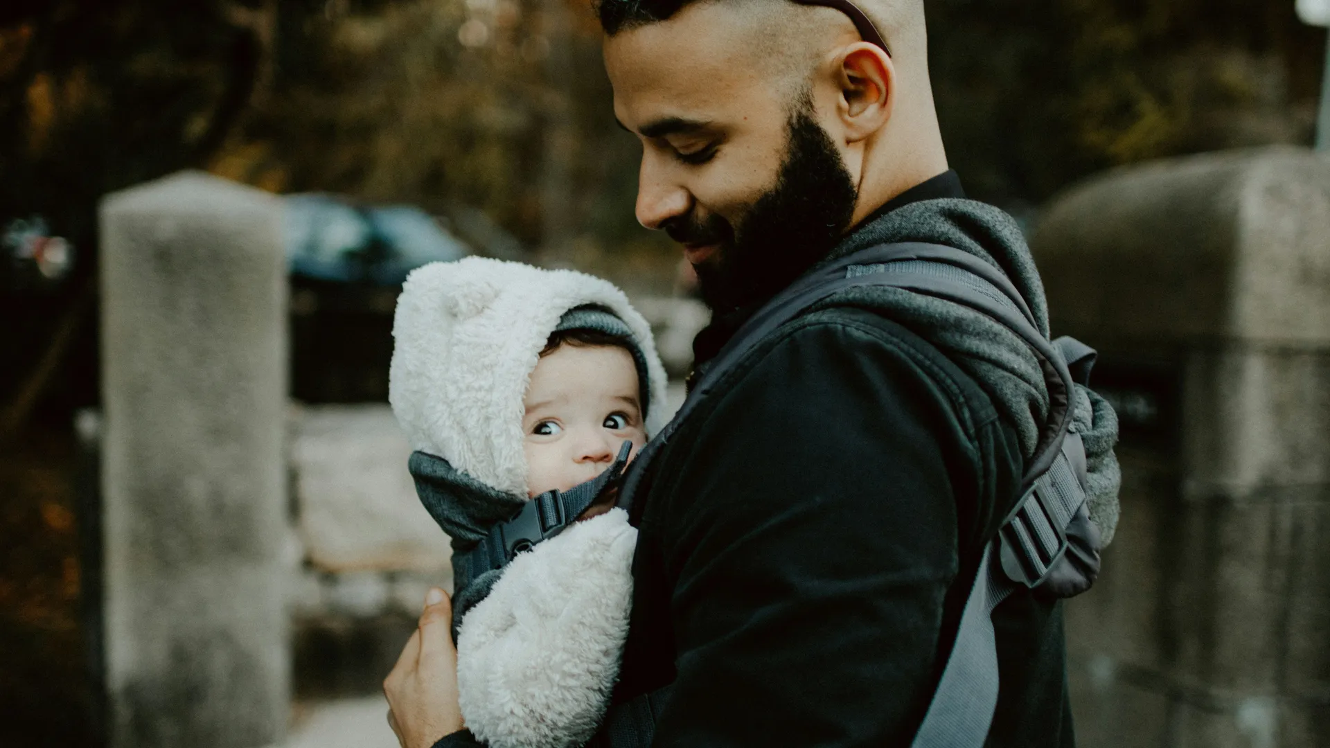 Smiling man with a beard carrying a bundled-up baby in a front carrier on a chilly day; the baby looks to the side while snuggled in a fluffy hooded outfit.