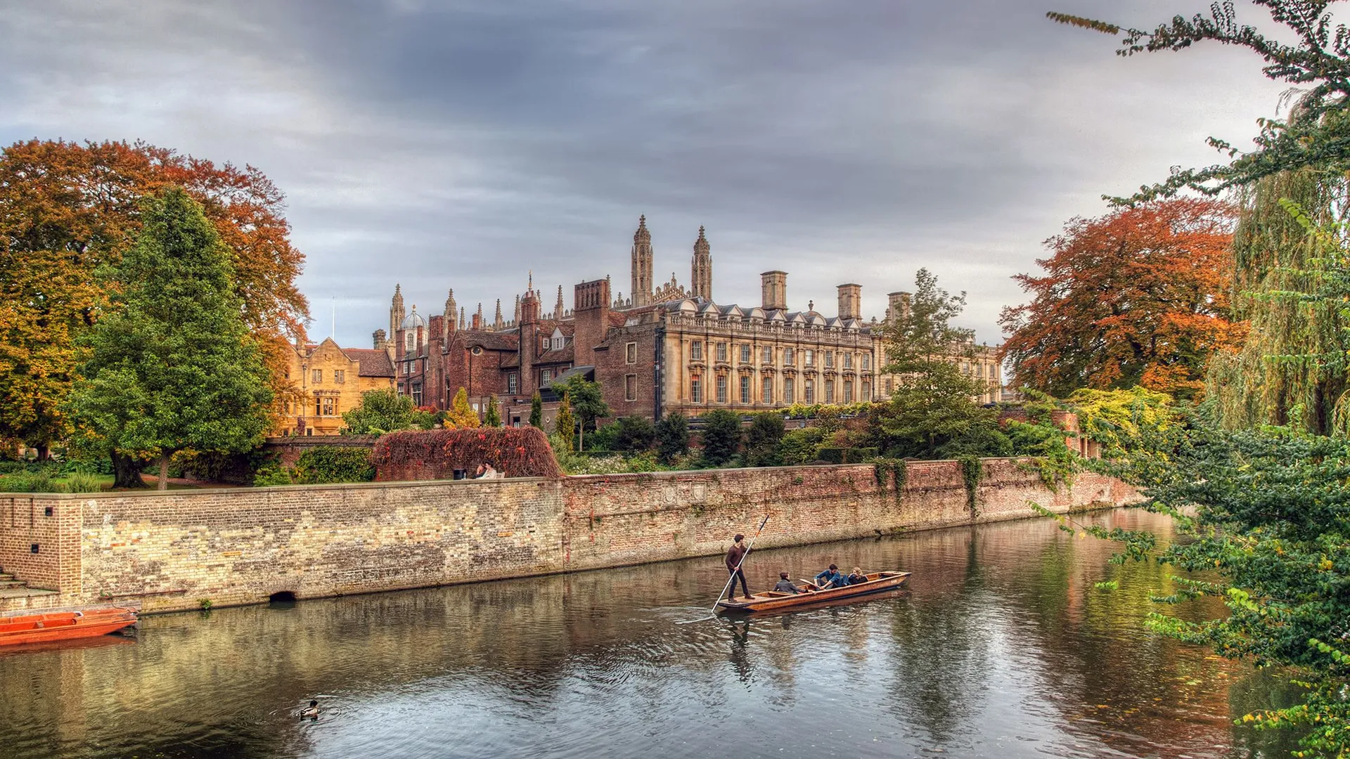 A scenic autumn view of Cambridge with colorful trees, historic university buildings, and people punting on the river.